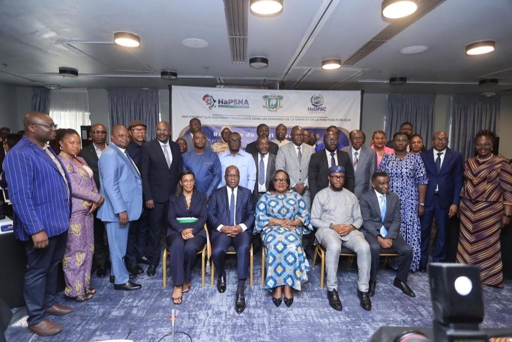 About thirty delegates in formal dress pose for a group photograph in a conference room, five seated in front. The backdrop carries the HaPSNA, Côte d’Ivoire and HeDPAC emblems above the words “Partenariat Sud-Sud pour l’excellence dans les domaines de la santé et de la fonction publique”.